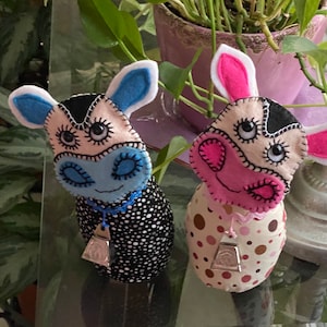 May include: Two handmade plush cow toys with black and white spots. One cow has blue and white ears and a blue collar with a silver bell. The other cow has pink and white ears and a pink collar with a silver bell.