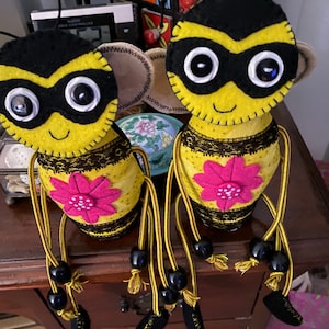 May include: Two felt bee figurines with black eyes, black masks, and yellow bodies. Each bee has a pink flower on its abdomen and black and yellow string legs with black beads.