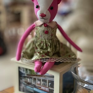 May include: A pink and white fabric doll with a green floral dress and white lace trim sitting on a white box with a picture of a trolley car. The box is on a table with a book titled "The Little Book of Hygge" and a glass jar with cookies.