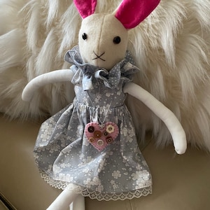 May include: A white bunny doll wearing a gray dress with white flowers and a pink heart-shaped button necklace. The doll has pink ears and black eyes.
