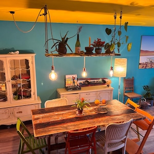 Könnte beinhalten: Ein Esszimmer mit einem rustikalen Holztisch und verschiedenen Stühlen. Über dem Tisch hängt ein Holzregal mit Pflanzen und hängenden Glühbirnen. Die Wände sind türkis und die Decke ist gelb.