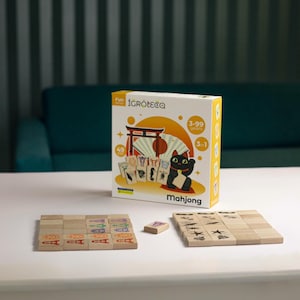 May include: A boxed Mahjong game with a Japanese theme, featuring a lucky cat illustration. The box displays the game's name and age range. Two sets of wooden tiles are arranged on a white surface, ready for play.