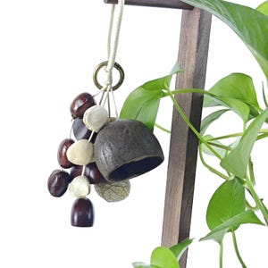 Pode incluir: Um ornamento suspenso com um cacho de sementes polidas castanho-escuras, sementes planas de cor creme e um sino de casca de coco castanho-escuro. Os componentes estão suspensos num anel de latão por um cordão de algodão entrançado cru.