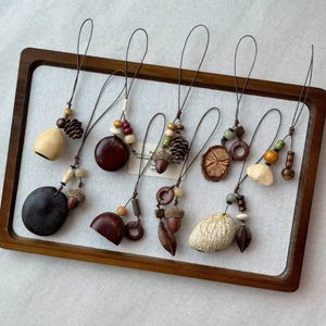 May include: A collection of seven hanging ornaments featuring natural elements including wooden beads, small pinecones, acorn caps, and polished dark brown seeds. Each ornament is suspended by a thin dark cord, showcasing a variety of textures and earthy tones.