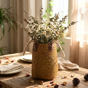 May include: A woven seagrass basket with two jute rope handles and brown leather accents, filled with a bouquet of dried eucalyptus branches and small white daisy flowers. The basket features a textured, cross-hatched weave pattern.