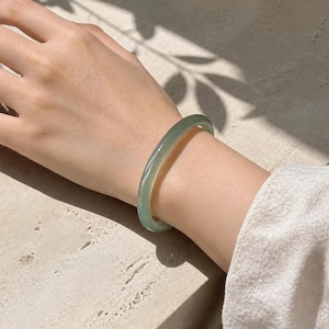 Pode incluir: Uma pulseira tipo bracelete lisa e translúcida de cor verde-pálido com acabamento polido. O acessório circular apresenta um perfil arredondado consistente e uma superfície suave que reflete a luz, exibindo variações subtis no tom verde.