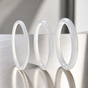 Può includere: Tre bracciali circolari di colore bianco traslucido e superficie liscia, disposti in fila. Ogni bracciale presenta una finitura lucida e arrotondata con uno spessore costante. I bracciali variano leggermente nel diametro e sono verticali.