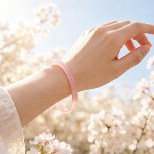 May include: A smooth, translucent light pink bangle bracelet with a rounded profile. The bracelet is worn on a wrist, showcasing its solid, polished surface and consistent pale pink color throughout the entire circular band design.