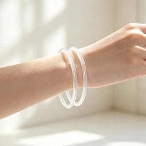 Può includere: Due bracciali rigidi bianchi, lisci e traslucidi, con profilo arrotondato. Questi accessori circolari presentano un aspetto chiaro simile al vetro e un design semplice e privo di decorazioni, indossati comodamente al polso come coppia.