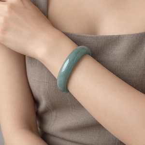 May include: A smooth, solid, rounded bangle bracelet in a muted sage green color. The bangle features a polished, glossy finish with subtle variations in tone throughout the stone, creating a continuous, seamless circular band design.