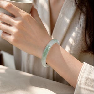 以下が含まれることがあります： 淡い緑色の翡翠で作られた滑らかで磨き上げられたバングルブレスレット。白と濃い緑色の斑点状のインクルージョンが特徴です。円形のジュエリーは丸みを帯びた断面で、光を反射する光沢のある表面仕上げが施されています。