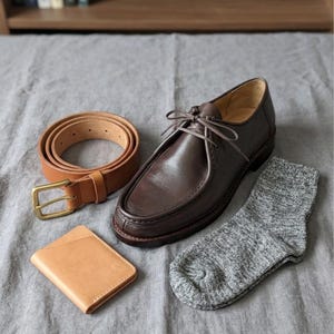 May include: A collection of men's accessories including a brown leather belt with a brass buckle, a pair of brown leather shoes, a light brown leather wallet, and a pair of gray socks. The items are arranged on a gray fabric surface.