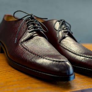 May include: A pair of burgundy leather dress shoes with black laces. The shoes have a textured surface and a classic design, suitable for formal occasions. The shoes are displayed on a wooden surface.