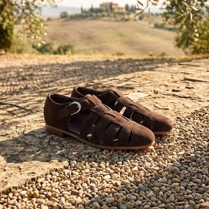 May include: A pair of dark brown suede leather fisherman sandals featuring a closed toe, multiple woven straps across the upper, and a side buckle closure. The footwear has a flat, low-profile sole and a smooth, matte texture throughout the exterior.