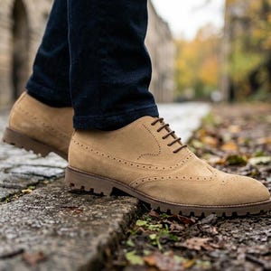 Peut inclure: Chaussures brogues en daim beige avec lacets marron foncé et semelle robuste. Les chaussures présentent des perforations décoratives et sont portées avec un jean bleu foncé. Image prise en extérieur sur une rue pavée humide.