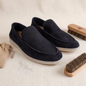 May include: A pair of dark navy blue suede slip-on loafers featuring a moccasin-style stitched toe seam and a light beige rubber sole. The shoes are accompanied by two wooden brushes with dark bristles and a small tan burlap drawstring pouch.