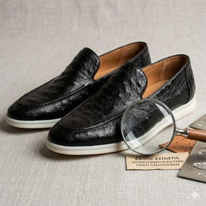 May include: A pair of black leather loafers with a textured ostrich emboss pattern. The shoes have a white sole and a brown interior. A magnifying glass and a card with text are next to the shoes.