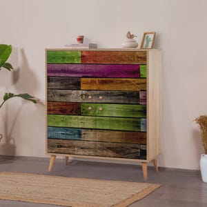 May include: A wooden chest of drawers with a colourful, rustic design. The front features a patchwork of painted wooden planks in various colours, including green, purple, and brown. The chest has four drawers and light-coloured wooden legs.