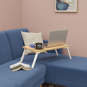 May include: A light wood-effect laptop table with white folding legs. A laptop, coffee cup, mouse, and cushion rest on the table, which is placed on a blue sofa. An open book is on the sofa. The background is a pink wall with a framed picture.