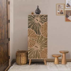 May include: A tall, wooden cabinet with a botanical design in shades of green and tan. The cabinet has a light wood top and legs. A decorative lantern and small wooden sphere sit on top. A woven basket and two small wooden stools are in front.