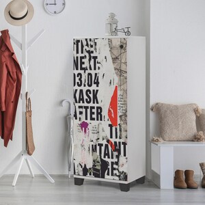 May include: A white cabinet with a collage design featuring text and abstract shapes. The cabinet has black feet and is placed against a white wall. A coat rack, umbrella, and bench are also visible.