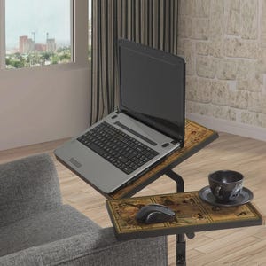 Op de afbeelding: Een zilveren laptop staat op een in hoogte verstelbare tafel met twee niveaus. De bovenste laag houdt de laptop vast, terwijl de onderste laag een muis en een theekopje met schotel heeft. De tafel heeft een decoratief kaartontwerp.