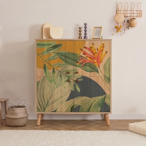May include: A wooden cabinet with a botanical design featuring large green leaves and a vibrant orange and yellow flower. The cabinet has four doors, wooden knobs, and angled legs. The background is a light beige.