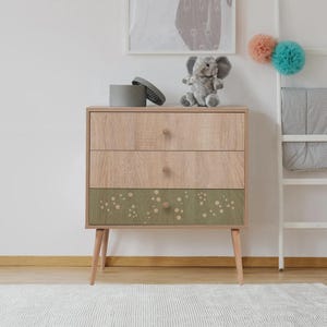 Puede incluir: Cómoda de madera de tres cajones con acabado de veta de madera clara. El cajón inferior es de color verde oliva con un estampado de puntos blancos. Tiene pomos redondos de madera y patas anguladas. Un elefante de peluche gris encima.