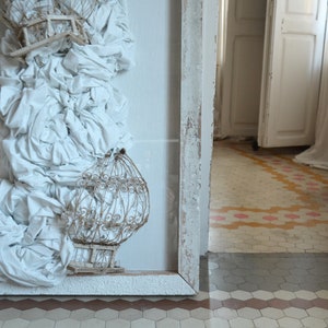 Può includere: Un'opera d'arte incorniciata in bianco che presenta una scultura in tessuto bianco con una gabbia per uccelli nell'angolo in basso a destra. L'opera d'arte è appesa a una parete bianca con un pavimento piastrellato sullo sfondo.