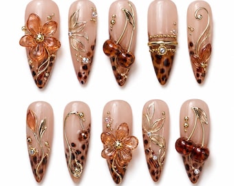 May include: A set of ten almond-shaped press-on nails featuring a nude base with brown leopard print tips. The designs include three-dimensional amber-colored flowers, gold metallic leaf accents, small clear rhinestones, and glossy brown cherry charms.