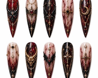 May include: A collection of ten long, pointed artificial nails with intricate gothic designs. Each nail features a base color of red, black, or cream, adorned with gold chains, stars, crosses, and devil or bat motifs. The designs are detailed and ornate.