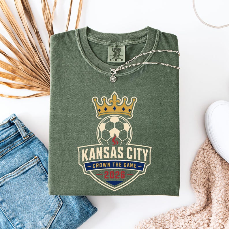 May include: Olive green t-shirt with a soccer-themed graphic. The graphic includes a soccer ball, a gold crown, and the text "Kansas City - Crown the Game - 2026" in a shield design. A silver necklace is draped across the shirt.