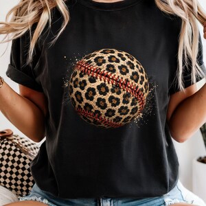 May include: A black short-sleeve t-shirt featuring a central graphic of a baseball with a brown and black leopard print pattern and prominent red stitching. The design includes subtle paint splatter effects surrounding the baseball.