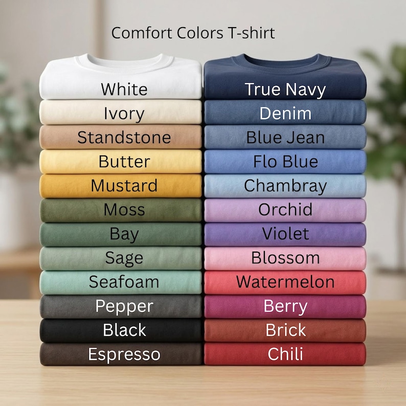 May include: Stacks of Comfort Colors T-shirts in various colors, including white, navy, and black. The image displays a range of hues, from pastels to darker shades, with each shirt neatly folded and labeled with its color name.