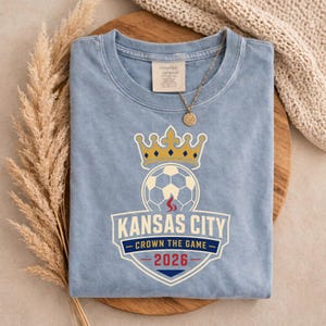 May include: A light blue t-shirt with a soccer ball and crown graphic. The graphic includes the text "Kansas City Crown the Game 2026". The shirt is folded and has a tag at the neckline. A gold necklace with a pendant is on top of the shirt.
