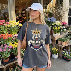 May include: A charcoal gray t-shirt featuring a soccer ball with a crown above it and the words "Kansas City" and "Crown the Game 2023" in a shield design. The shirt is worn with a white baseball cap and denim shorts.