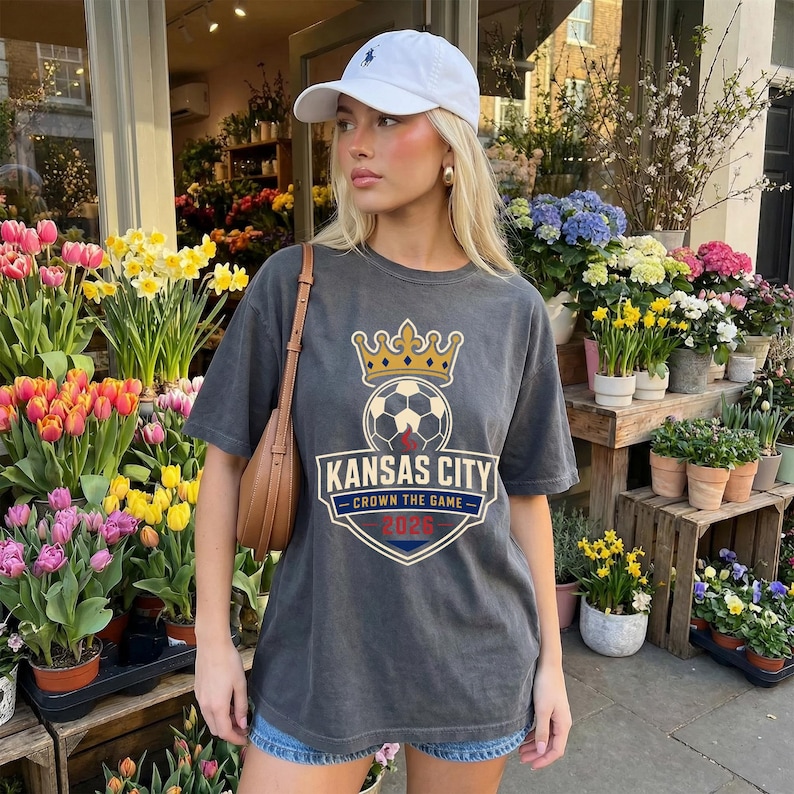 May include: A charcoal gray t-shirt featuring a soccer ball with a crown above it and the words "Kansas City" and "Crown the Game 2023" in a shield design. The shirt is worn with a white baseball cap and denim shorts.