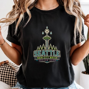 May include: Black t-shirt featuring a graphic design for the Seattle Rain City Match Soccer 2026. The design includes the Space Needle, a soccer ball, and the text "SEATTLE" in a teal and white color scheme. The shirt has rolled-up sleeves.