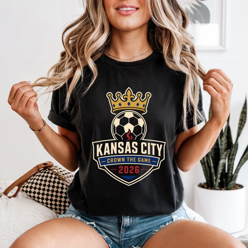 May include: Black t-shirt featuring a soccer ball with a crown, the words "Kansas City" and "Crown the Game 2026". The design includes red, blue, and gold accents. The shirt is made of a soft material.