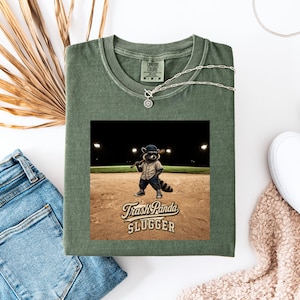 Può includere: T-shirt verde oliva con grafica di un procione in uniforme da baseball con una mazza, con la scritta "Trash Panda Slugger". La maglietta è abbinata a una collana d'argento e jeans blu.