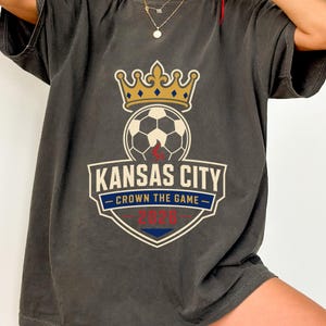 May include: Dark gray t-shirt featuring a soccer ball topped with a gold crown. The shirt displays the text "Kansas City - Crown the Game - 2026" in a shield design. The shirt is a comfortable, casual style.