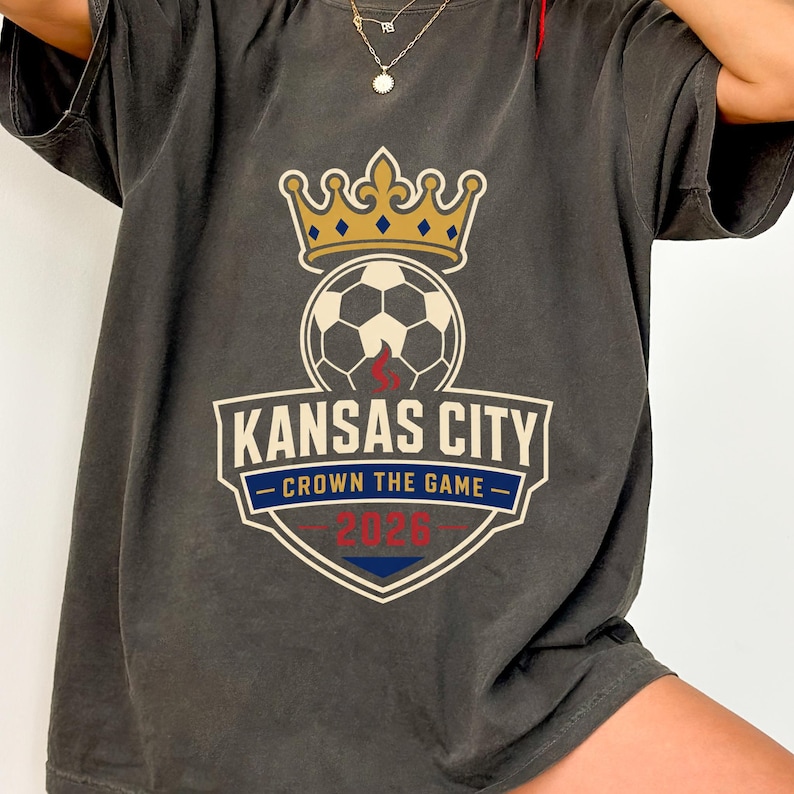 May include: Dark gray t-shirt featuring a soccer ball topped with a gold crown. The shirt displays the text "Kansas City - Crown the Game - 2026" in a shield design. The shirt is a comfortable, casual style.