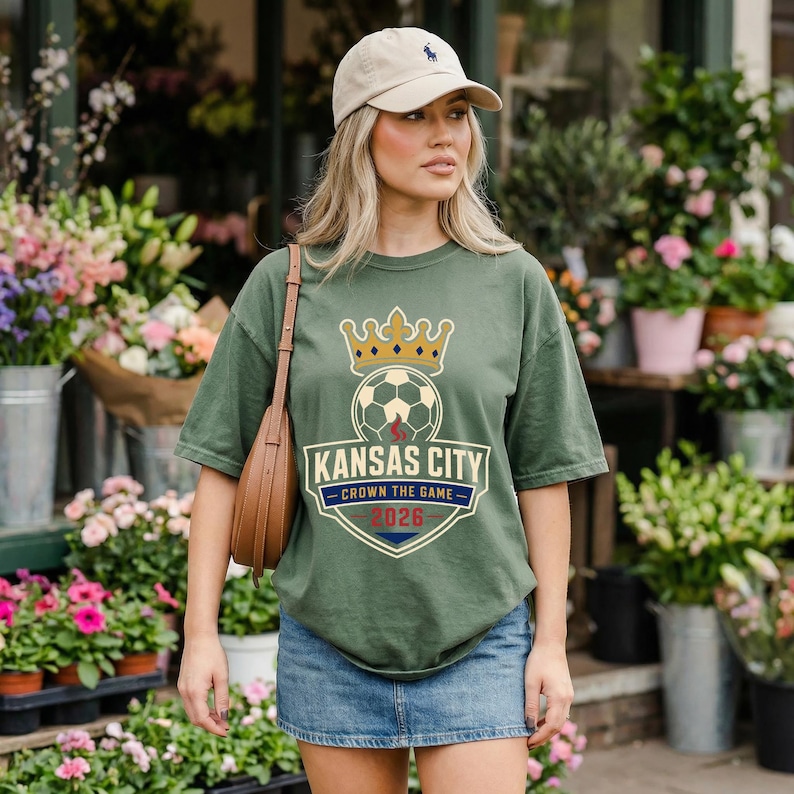 May include: Olive green t-shirt with a soccer-themed graphic. The design features a soccer ball with a crown above it and the words "Kansas City Crown the Game 2026" in a shield shape. The shirt is worn with a denim skirt and a tan baseball cap.