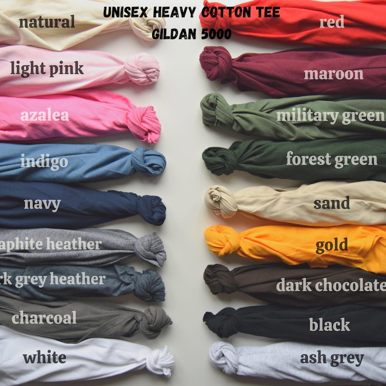 May include: A display of folded unisex heavy cotton tees in various colors, including red, navy, and black. The image shows the Gildan 5000 model, with color names such as "natural," "light pink," and "ash grey" labeled next to each folded shirt.