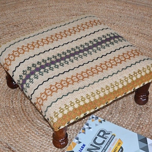 May include: A small, square footstool with a patterned fabric top in beige, orange, purple, and black. The stool has four short, dark wooden legs. The stool is on a woven jute rug.