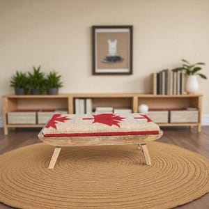 Peut inclure: Un banc bas en bois doté d'un coussin rectangulaire avec une base en tissu beige et un motif répétitif de feuilles d'érable rouges. Le banc possède quatre pieds en bois clair inclinés et une finition en grain de bois naturel.