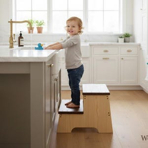 Può includere: Uno sgabello in legno chiaro a due gradini. Un bambino piccolo è in piedi sullo sgabello, raggiungendo un piano di lavoro della cucina. La cucina presenta mobili bianchi, un piano di lavoro in marmo e un rubinetto dorato.