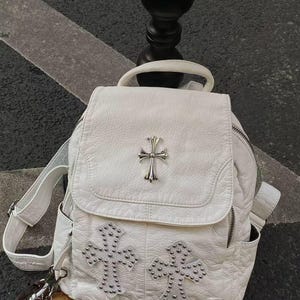 Pode incluir: Uma mochila branca texturizada com um emblema de cruz em metal prateado na aba superior e dois grandes padrões em forma de cruz decorados com pequenas tachas prateadas no bolso frontal. A mochila inclui uma alça superior e bolsos laterais.