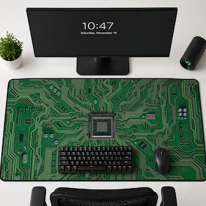 Könnte beinhalten: Ein großes rechteckiges grünes und schwarzes Computer-Mauspad mit Leiterplatten-Design. Eine schwarze Tastatur und Maus befinden sich auf dem Pad. Ein Computermonitor zeigt die Uhrzeit und das Datum an. Eine kleine Pflanze und ein schwarzer Zylinder sind ebenfalls vorhanden.