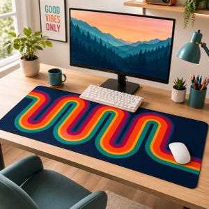 May include: A rectangular desk mat featuring a dark blue background with a repeating, wavy, multi-coloured stripe pattern in shades of orange, yellow, and teal. The mat is positioned on a wooden desk surface beneath a computer monitor and keyboard.